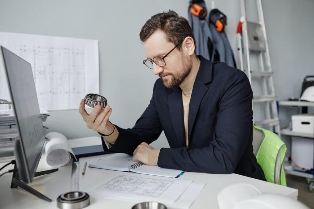 Bearded mechanical engineer holding machine part at workplace